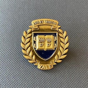 May include: A circular brass-toned metal pin featuring the Yale University emblem. The design includes a central shield with a blue background, an open book with Hebrew text, and laurel leaves. Banners read LUX ET VERITAS at the top and YALE at the bottom.