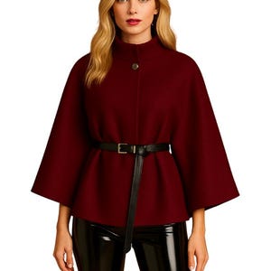 May include: A burgundy cape with a high collar and a single button closure. The cape features wide sleeves and is cinched at the waist with a black belt. The model is wearing black, shiny pants.