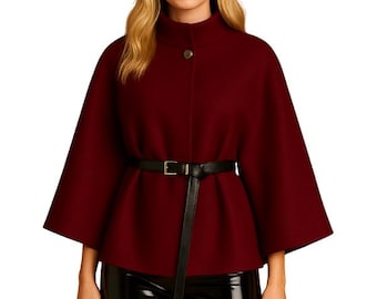 May include: A burgundy cape with a high collar and a single button closure. The cape features wide sleeves and is cinched at the waist with a black belt. The model is wearing black, shiny pants.