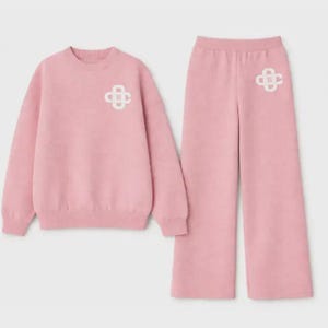 May include: A matching pink sweatshirt and wide-leg pants set. The sweatshirt and pants feature a white emblem on the front. The set is made of a soft, comfortable material, perfect for casual wear.