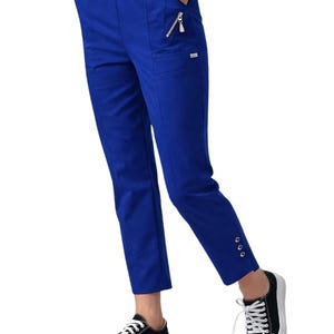 May include: Royal blue trousers with an elasticated waistband, side pockets, and a zip detail. The trousers have a straight leg cut and are cuffed at the ankle with three gold-coloured buttons. The person is wearing black and white trainers.