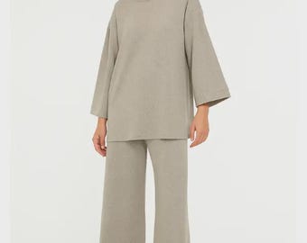 May include: A two-piece set of casual wear in a neutral beige color. The set includes a long-sleeved, oversized top and wide-leg pants. The model is wearing the outfit with beige sandals. The outfit is made of a soft, textured fabric.
