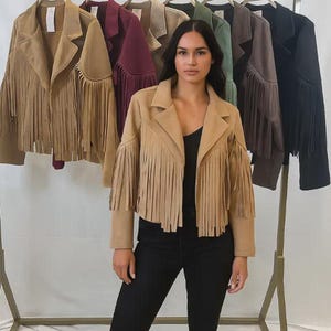 May include: A woman wearing a tan suede fringe jacket, posing in front of a rail of jackets in various colours. The jackets have fringe detailing on the sleeves and hem. Other colours include burgundy, olive green, and black.