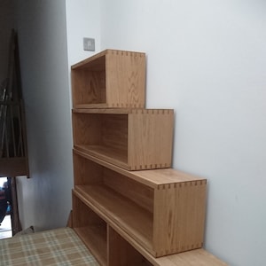 May include: A set of five wooden shelves stacked in a stair-step pattern. The shelves are made of light brown wood and have a natural finish. The shelves are mounted on a wall and are empty.