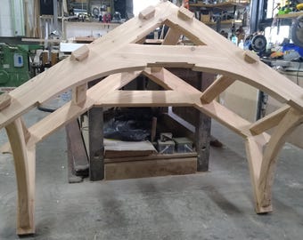 May include: A wooden archway structure with a triangular top, constructed from unfinished wood beams. The archway is supported by two curved beams on each side.