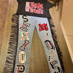 May include: Blue denim jeans with black fringe and the word "SENIOR" spelled out in black and white letters with red hearts. The jeans have a patch on the right leg that says "They Not Like Us 2025" in red and white letters.