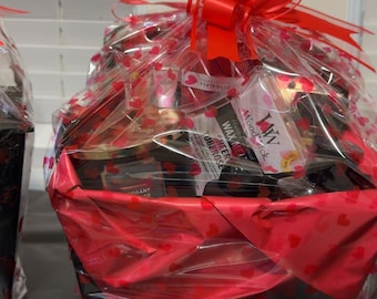 May include: A gift basket wrapped in clear cellophane with red heart patterns, topped with a large red ribbon bow. The basket contains various items including a WoodWick wax melt labeled Sheer Tuberose and other assorted packaged goods.