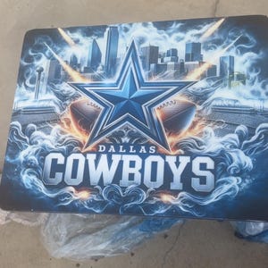 May include: A rectangular table featuring a Dallas Cowboys design. The design includes a large blue star, footballs, a cityscape, and the words "DALLAS COWBOYS" in white. The overall color scheme is blue and white.