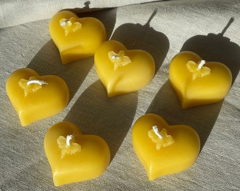May include: Six yellow heart-shaped candles with a small white butterfly design on each. The candles are arranged on a light beige fabric surface.