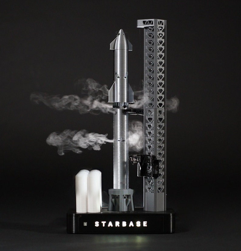 May include: A silver model rocket on a launchpad with smoke coming from the base. The launchpad has a black base with the text "STARBASE" printed on it.