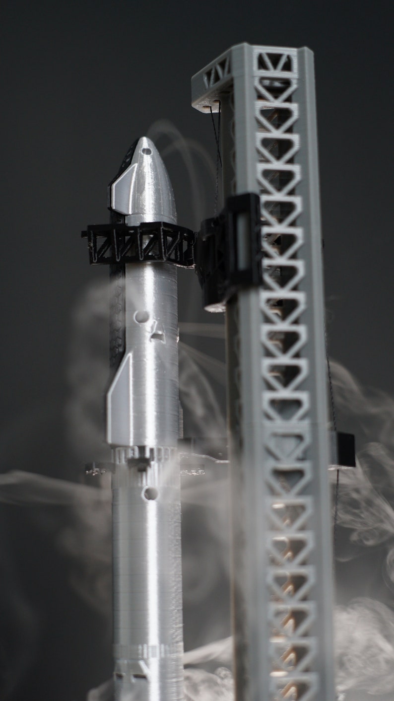 May include: A silver model rocket on a launchpad with smoke coming from the base. The rocket is attached to the launchpad with a black metal structure.