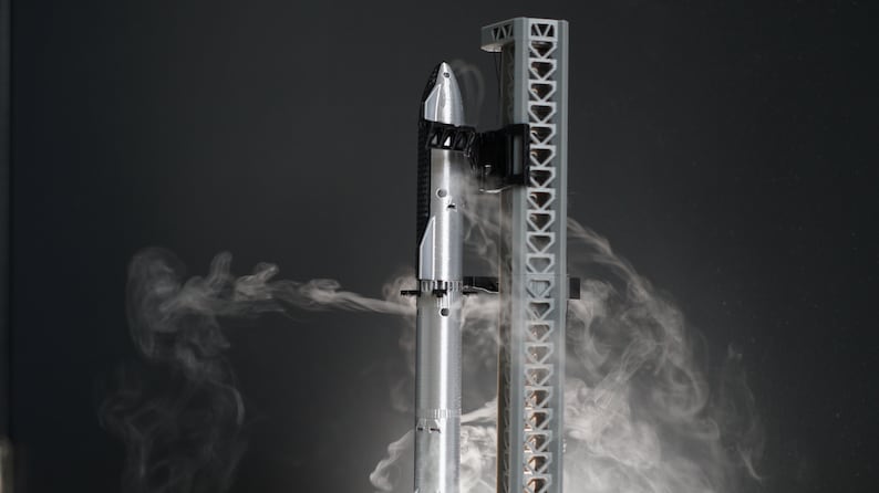 May include: A silver model rocket with black accents is mounted on a grey launch tower. Smoke is billowing around the rocket, creating a dramatic effect.