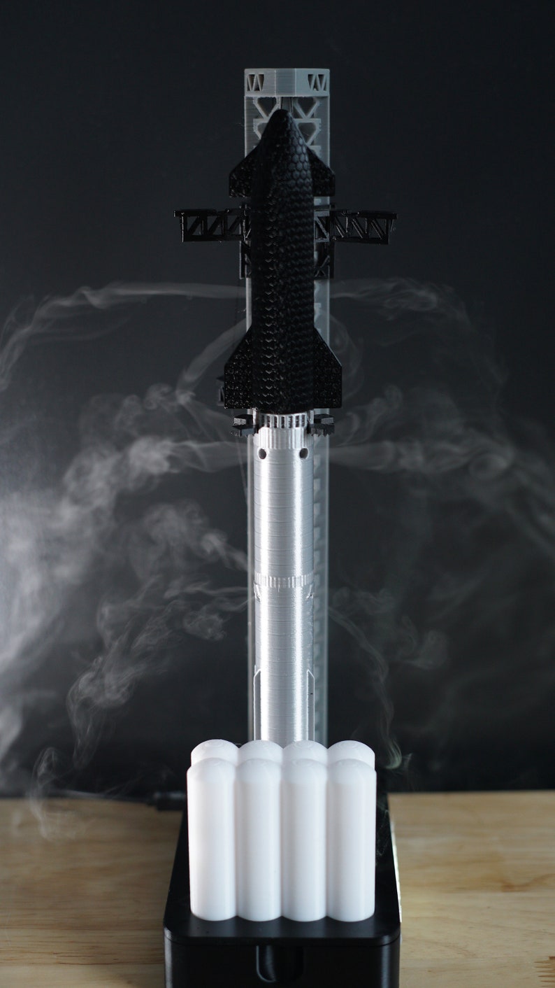 May include: A 3D printed model of a rocket ship with smoke emanating from the bottom. The rocket is black and white and is standing on a white base. The base has a black platform with a small opening at the front.