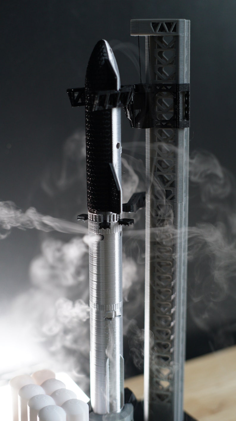 May include: A 3D printed model of a rocket on a launchpad with smoke coming from the base. The rocket is silver and black, and the launchpad is grey.