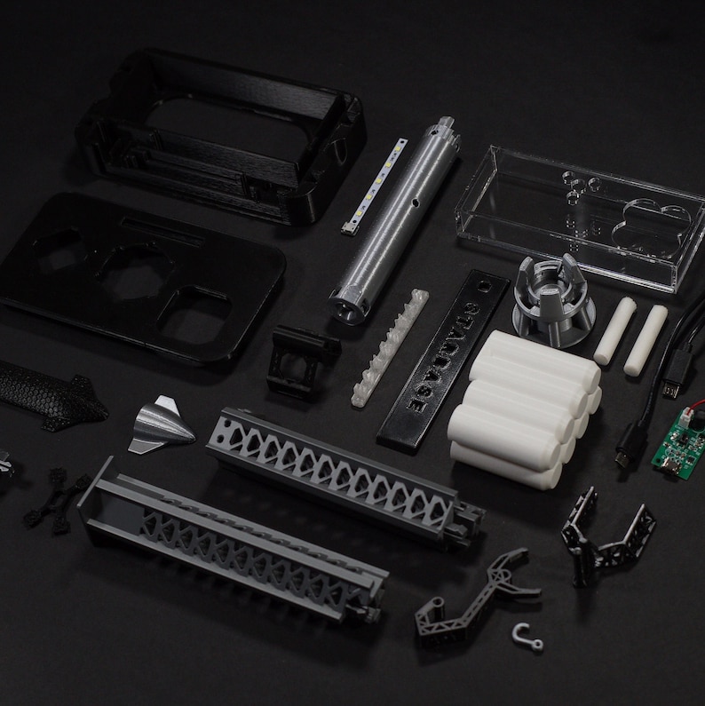 May include: A collection of 3D printed parts for a DIY project. The parts are black, grey, and white. The parts include a small rocket, a long tube with a grid pattern, a small box, and a clear plastic box. The parts are arranged on a black surface. The text "SABOTAGE" is printed on a black rectangular piece of plastic.