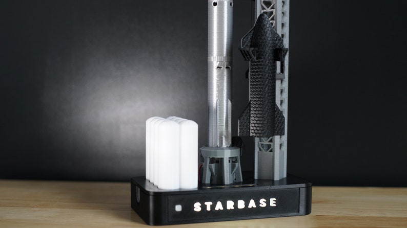 May include: A black and grey model rocket on a stand with the word "STARBASE" printed on the base. The rocket is attached to a grey launch tower. There are four white cylindrical objects on the base.