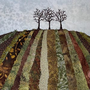 May include: Textile artwork showing three bare trees on a hill. The hill is made of vertical fabric strips in earth tones: brown, green, and beige. The sky is pale blue with subtle cloud-like patterns. A landscape-themed textile art piece.