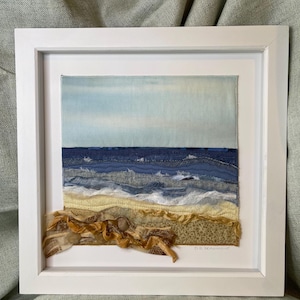 Pode incluir: Uma peça de arte têxtil enquadrada que retrata uma paisagem marítima com água azul, ondas brancas e uma praia de areia. A peça é assinada "D.L. Seamon".
