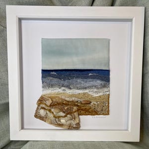 May include: A framed textile artwork depicting a seascape with blue water, white waves, and a sandy beach. The beach is decorated with brown and beige lace and white seashells. The artist's signature, "D.B. Beaumont", is visible in the lower right corner.