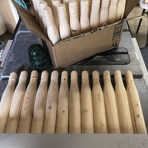 May include: A collection of unfinished wooden bottle-shaped objects, some arranged in a cardboard box, others laid out on a surface. The objects have a textured top and a smooth, tapered body, suggesting they are components for a craft project or decorative item.