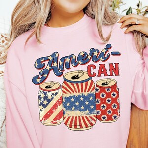 Puede incluir: Sudadera rosa con el texto "Ameri-Can" en una fuente vintage. Tres latas rojas, blancas y azules con estrellas y rayas están debajo del texto.