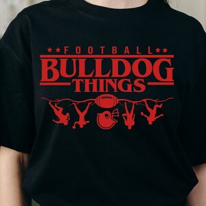 May include: A black t-shirt with a red graphic design that says "Football Bulldog Things" with a football and five stick figures hanging upside down. The design is inspired by the popular Netflix show "Stranger Things".