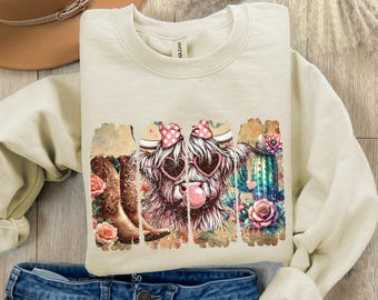 May include: A beige sweatshirt with a graphic print of a Highland cow wearing pink sunglasses and a pink bow. The cow is surrounded by flowers and a cactus. The graphic has a distressed look.