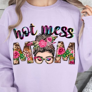 Può includere: Una felpa color lavanda con il testo "Not Mess Mom" in colori arcobaleno. Il testo è circondato da disegni floreali e con stampa leopardata. Un viso di donna con un chignon e fiori nei capelli è visibile al centro del testo.