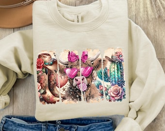 May include: A beige sweatshirt with a graphic print of a cow wearing pink sunglasses and a bow. The cow is surrounded by cacti, flowers, and cowboy boots.