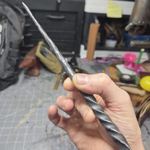 May include: A hand-forged metal wand with a spiraled handle and a pointed tip. The wand is a dark gray color and appears to be made of iron or steel. The wand is held in a person's hand.