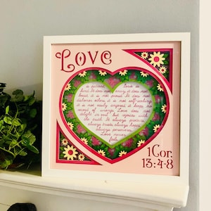 May include: A white frame with a pink background and a heart-shaped cutout. The heart is decorated with green leaves and pink flowers. The text "Love" is written in pink at the top of the frame. Inside the heart, a passage about love is written in cursive. The text "1Cor. 13:4-8" is written in pink at the bottom of the frame.