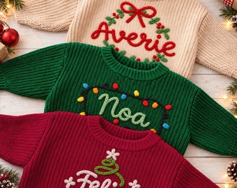 Könnte beinhalten: Drei gerippte Strickpullover in Beige, Grün und Rot. Der beige Pullover zeigt einen roten gestickten Kranz und den Namen Averie. Der grüne Pullover hat eine bunte Lichterkette und den Namen Noa. Der rote Pullover zeigt einen grünen Baum und den Namen Felix.