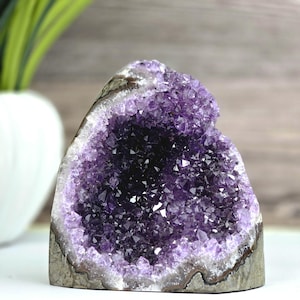 May include: A raw amethyst geode featuring a deep purple, crystalline interior with sharp, multifaceted points. The outer edge of the stone displays a natural, rough, greyish-brown rock formation that frames the vibrant violet crystal cluster.
