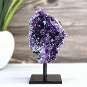 May include: A large, raw amethyst geode crystal cluster, showcasing deep purple hues and crystalline structures. The geode is mounted on a black metal stand with a square base, creating a striking display piece. The background is a blurred wooden surface.