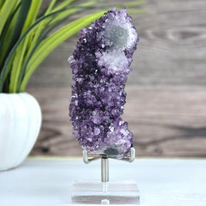 May include: A vertical amethyst crystal cluster featuring deep purple, faceted points and a white, crystalline center. The specimen is securely mounted on a silver-toned metal stand with a clear, square acrylic base for stable display.