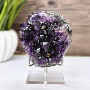 May include: A large, polished amethyst geode on a clear acrylic stand. The crystal features deep purple and black crystalline structures. The geode is displayed against a blurred background with a white vase and wooden paneling.