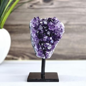 May include: A large, raw amethyst crystal cluster, showcasing deep purple hues and crystalline structures. The geode is mounted on a black metal stand with a square base, creating a striking display piece. The background features a wooden wall and a white vase.