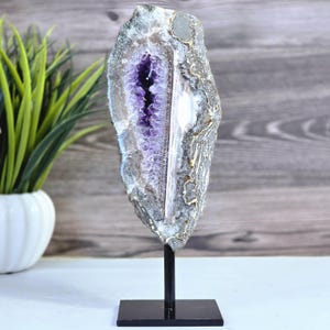 May include: A large amethyst geode slice, showcasing a deep purple crystal center within a gray and white banded exterior. The geode is mounted on a black metal stand, suitable for display as a decorative object.