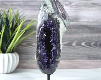 May include: A tall, vertical amethyst geode featuring a deep purple crystalline interior and a rough, greyish-white outer rock crust. The geode is securely mounted on a simple, black metal stand with a square base, displaying a natural formation.