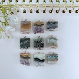 Puede incluir: Nueve colgantes de ágata de forma cuadrada con anillas de salto de plata. Los colgantes son de varios tonos de morado, verde, marrón y blanco. La ágata es translúcida y tiene un borde natural y rugoso.