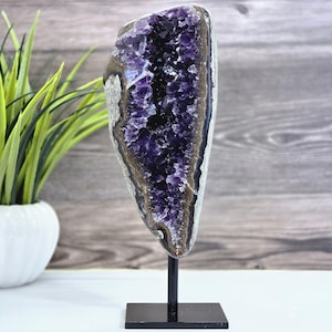 May include: A large, polished amethyst geode on a black metal stand. The geode features a deep purple crystalline interior, surrounded by a band of brown and gray rock. The stand has a square base and a vertical rod.