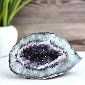 Può includere: Una geode aperta che rivela un gruppo di cristalli di ametista viola scuro. Il bordo esterno della geode è grigio chiaro con una consistenza cristallina. La geode è su una superficie bianca con uno sfondo in legno.
