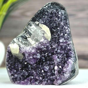 May include: A raw amethyst geode specimen featuring deep purple crystalline structures and a prominent cluster of translucent, pale yellow calcite crystals embedded within the upper section of the dark, jagged mineral formation.