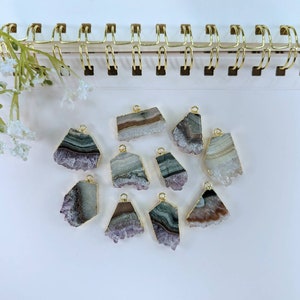 May include: Ten amethyst crystal pendants with gold-plated edges. The crystals are various shades of purple, white, and green. The pendants are arranged on a white surface.