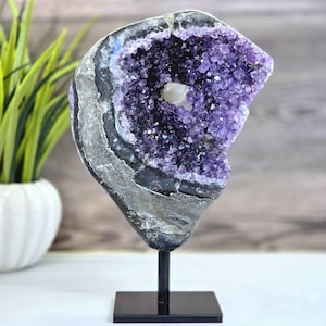 May include: A large amethyst geode crystal on a black metal stand. The geode features a deep purple interior with sparkling crystal formations and a white quartz center. The outer layers are banded with shades of gray and black.