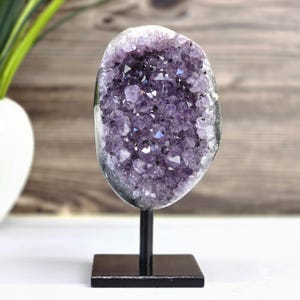 Könnte beinhalten: Ein großer, ovaler Amethyst-Geodenkristall auf einem schwarzen Metallständer. Die Geode hat eine tiefviolette Farbe mit funkelnden Kristallformationen. Der Ständer hat eine quadratische Basis und eine vertikale Stütze.