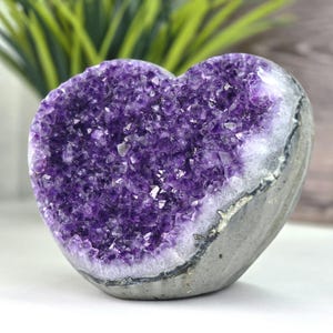 May include: A heart-shaped amethyst geode featuring deep purple crystalline structures on the front face, contrasted by a smooth, grey, natural stone exterior on the sides and base, showcasing the raw mineral formation in a polished heart shape.