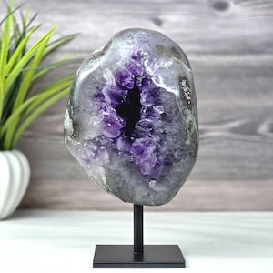 Könnte beinhalten: Eine polierte Amethyst-Geode mit einem tiefvioletten kristallinen Zentrum und einer grauen, strukturierten äußeren Gesteinsschale. Die Geode ist auf einem vertikalen schwarzen Metallstab montiert, der an einem quadratischen schwarzen Metallsockel befestigt ist.