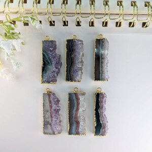 Puede incluir: Seis colgantes de cristal de amatista con ribete dorado. Los cristales son de diferentes tonos de púrpura y tienen una textura natural y rugosa.