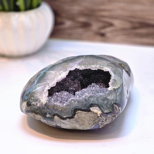 May include: A polished, oval-shaped geode with a hollow center filled with sparkling, dark purple crystals. The exterior is a mix of gray, green, and blue hues, with a smooth, glossy finish. The geode is a natural, decorative item.